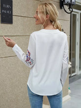 Load image into Gallery viewer, Woman wearing a white blouse with floral embroidery, standing outside a building.