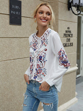 Load image into Gallery viewer, Woman wearing a white blouse with colorful embroidery and blue jeans standing in front of a building.