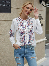 Load image into Gallery viewer, Woman wearing a white blouse with floral embroidery and ripped jeans, standing in front of a building.