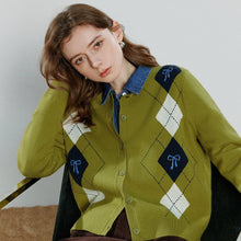 Load image into Gallery viewer, A woman wearing a green argyle cardigan with blue and white diamond patterns, layered over a blue collared shirt and paired with a brown skirt, sitting on a black chair. 
